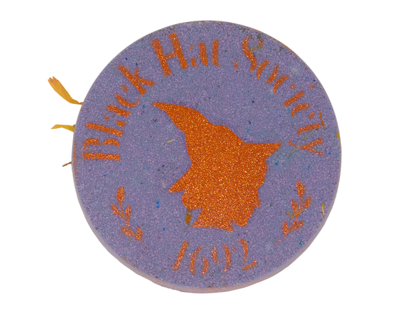 circular purple bath bomb with an autumn orange glitter airbrush of a witch's side profile surrounded by text that says black hat society 1692