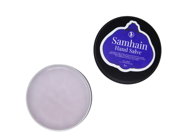 purple hand salve packaged in a 2 oz black tin with the lid removed to show the product. lid has a purple pumpkin shaped label that says Samhain Hand Salve with the product listed beneath it. 